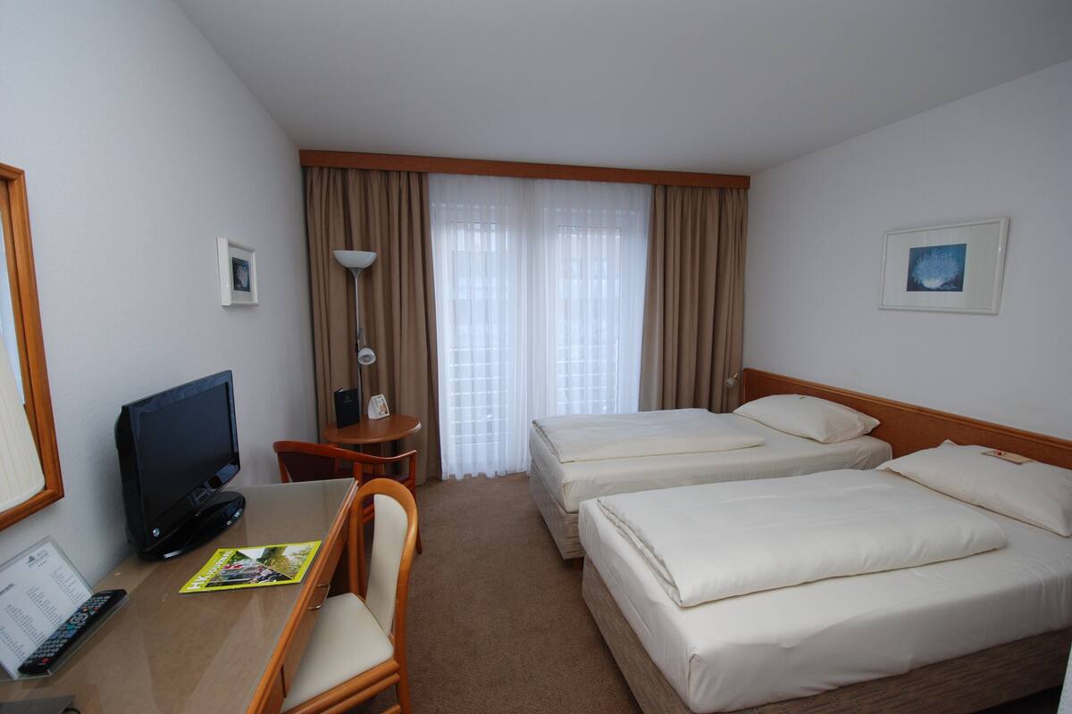 Een hotelkamer met twee bedden, een bureau met een computer, een stoel, een televisie en een raam met gordijnen.