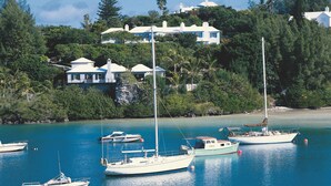 Un port de plaisance où sont amarrés plusieurs voiliers, entouré d’une végétation luxuriante et dominé par une grande maison blanche dotée de nombreuses cheminées.
