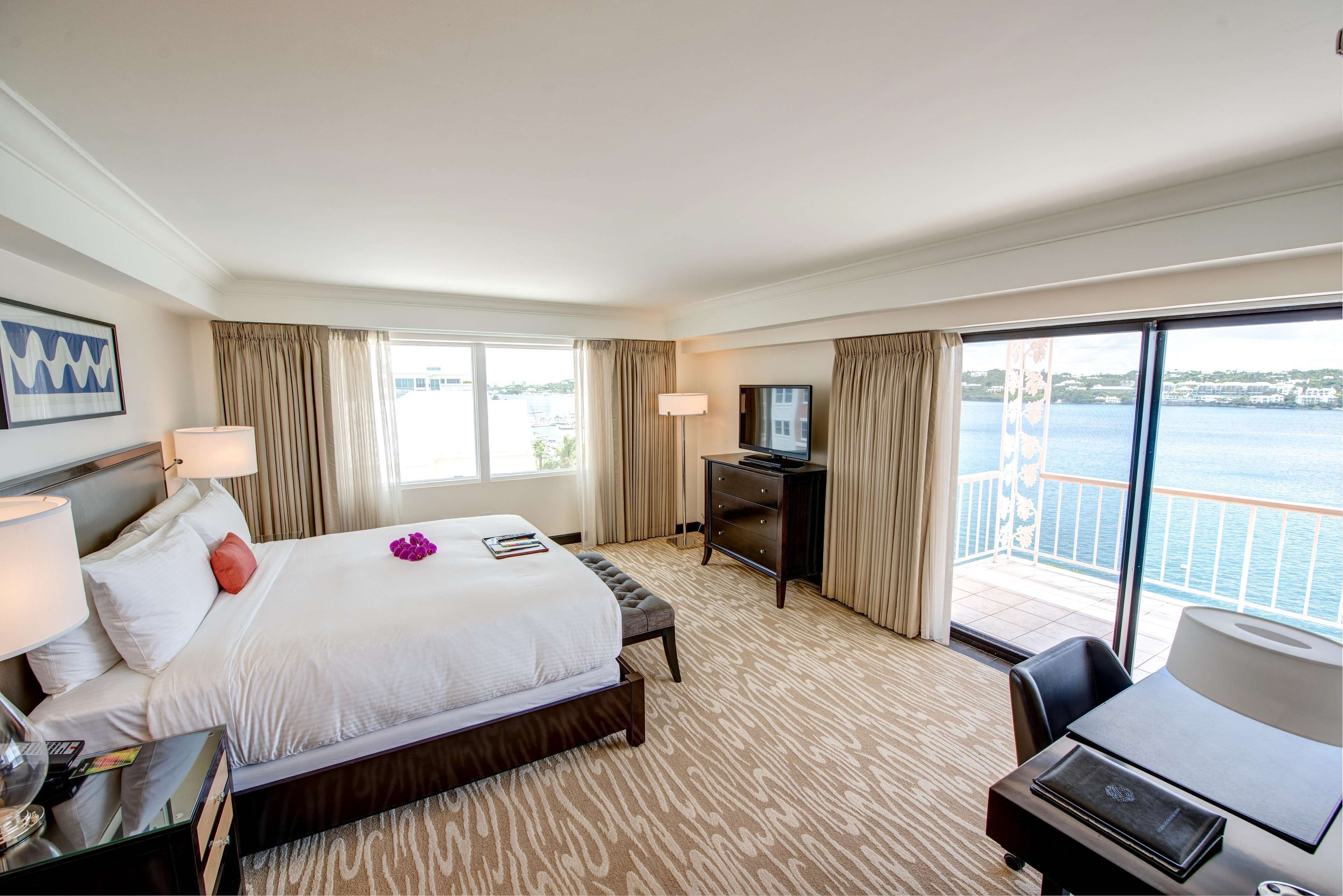A hotel room with a large bed, a desk, a TV, and a balcony with a view of the sea.