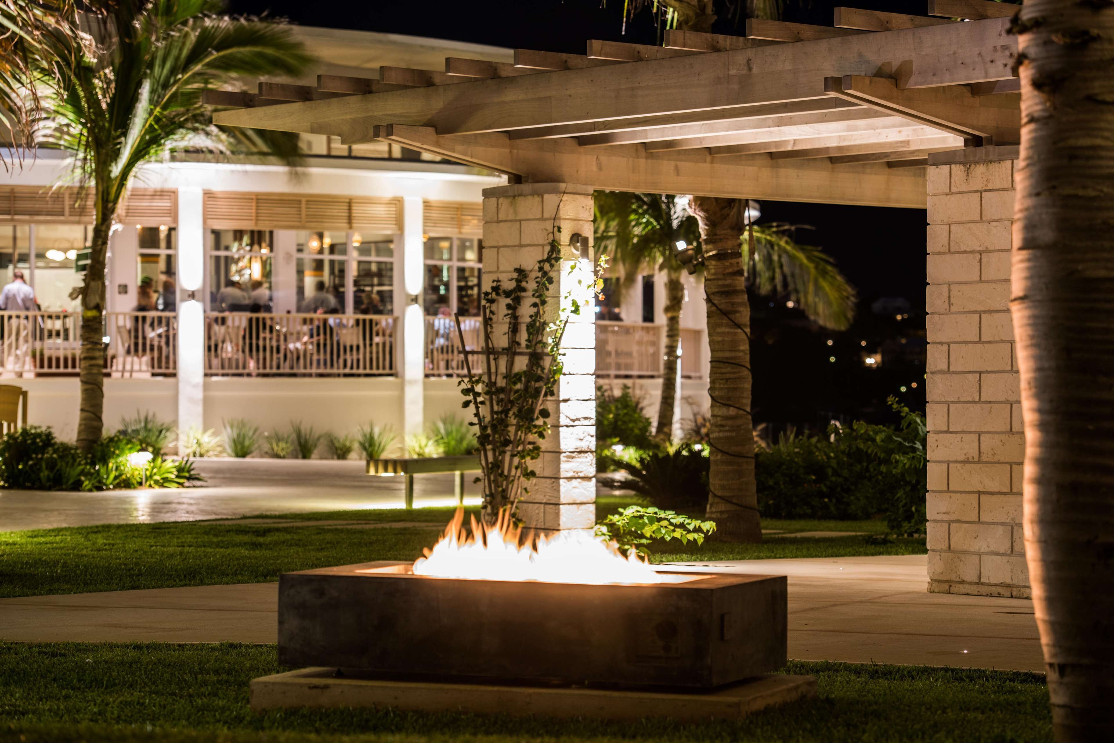 Una estructura de piedra con hogar al aire libre, rodeada por un patio iluminado con palmeras y un edificio al fondo.
