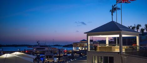 Un hotel frente al agua con una zona de estar al aire libre techada, un asta con una bandera y vistas al puerto deportivo.