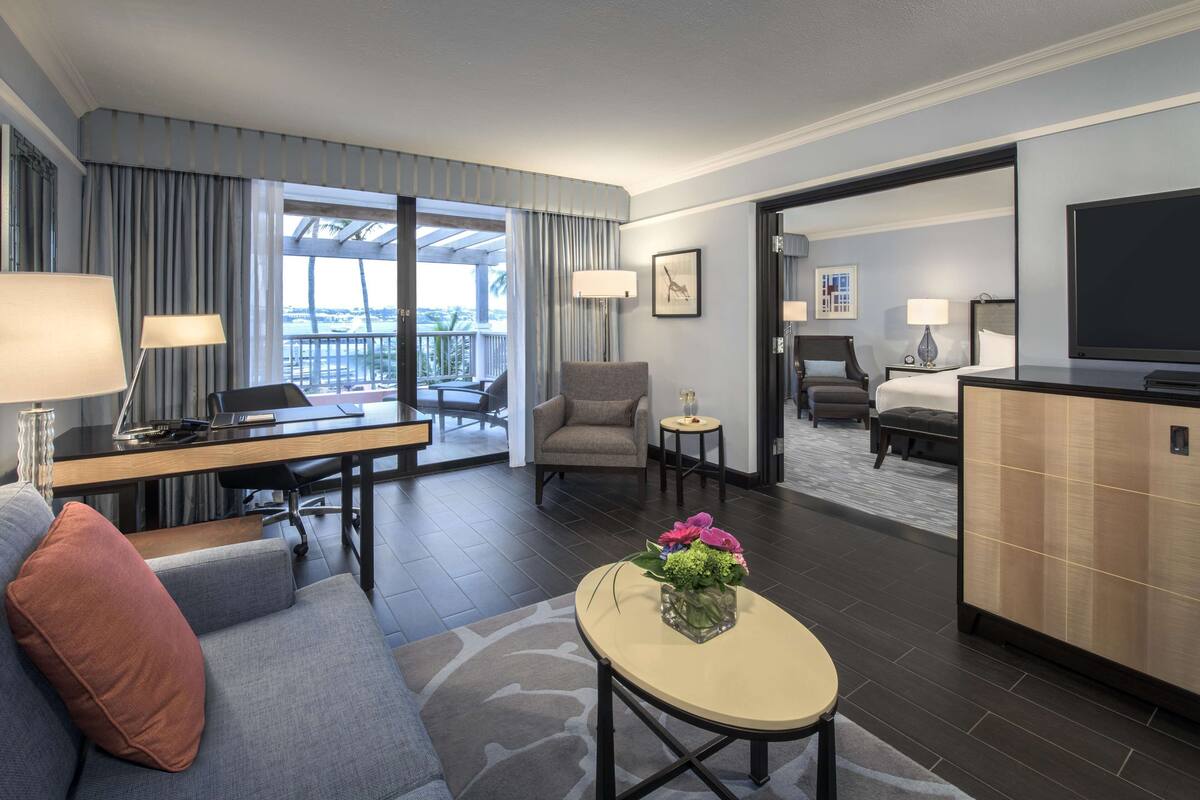 Ein Hotelzimmer mit Schreibtisch, Sofa, Ohrensessel und einem kleinen Tisch mit Blumen. Ein Balkon mit Vorhängen und Blick ins Freie ist ebenfalls vorhanden.