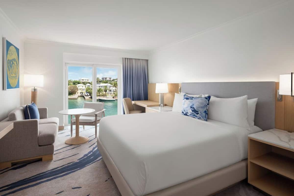 Ein Hotelzimmer mit einem großen Bett, einem Schreibtisch, einem Stuhl, einem kleinen runden Tisch und Blick auf den Pool sowie umliegende Gebäude.