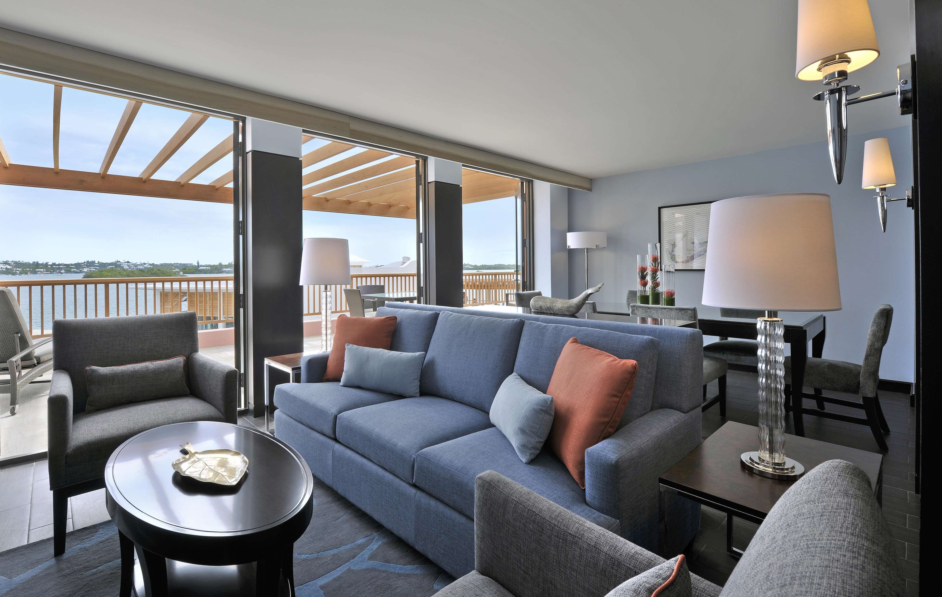 A modern hotel room with a balcony, a sofa set, a dining table, and a lamp.