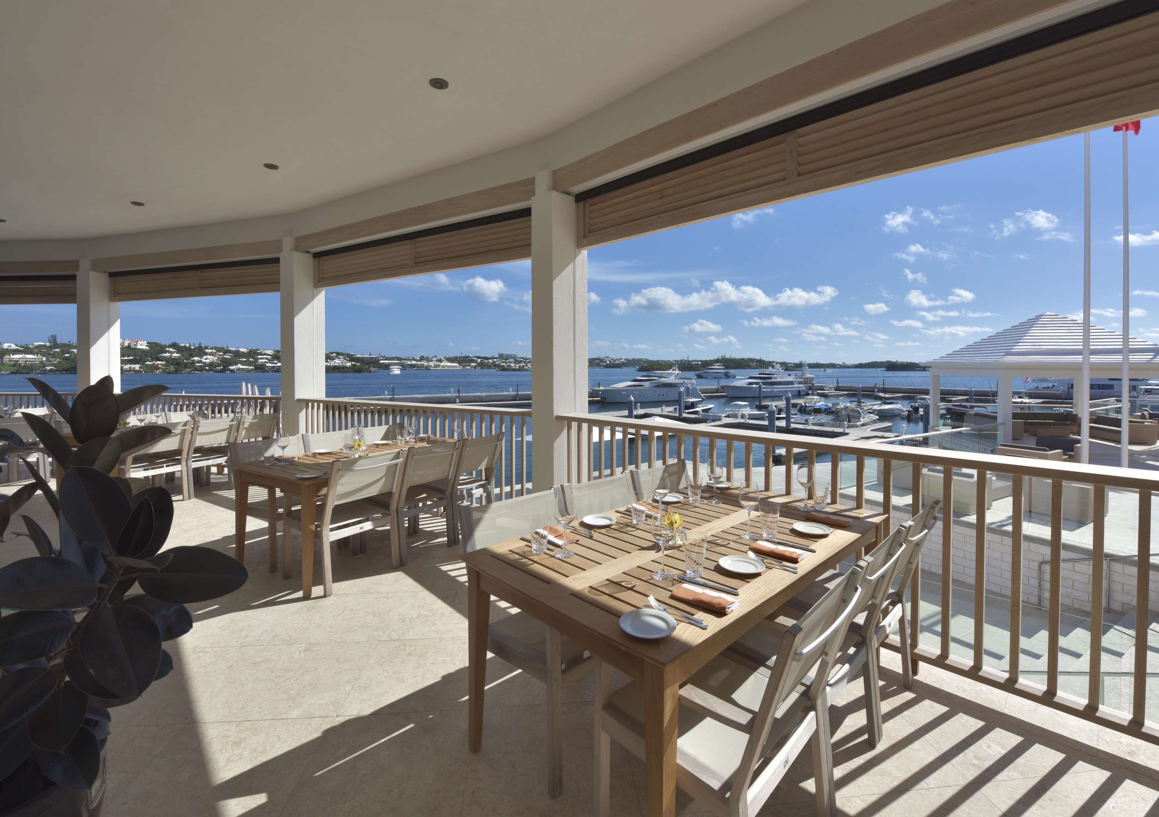 A dining area with tables set for two, overlooking a marina with boats and a distant shoreline.