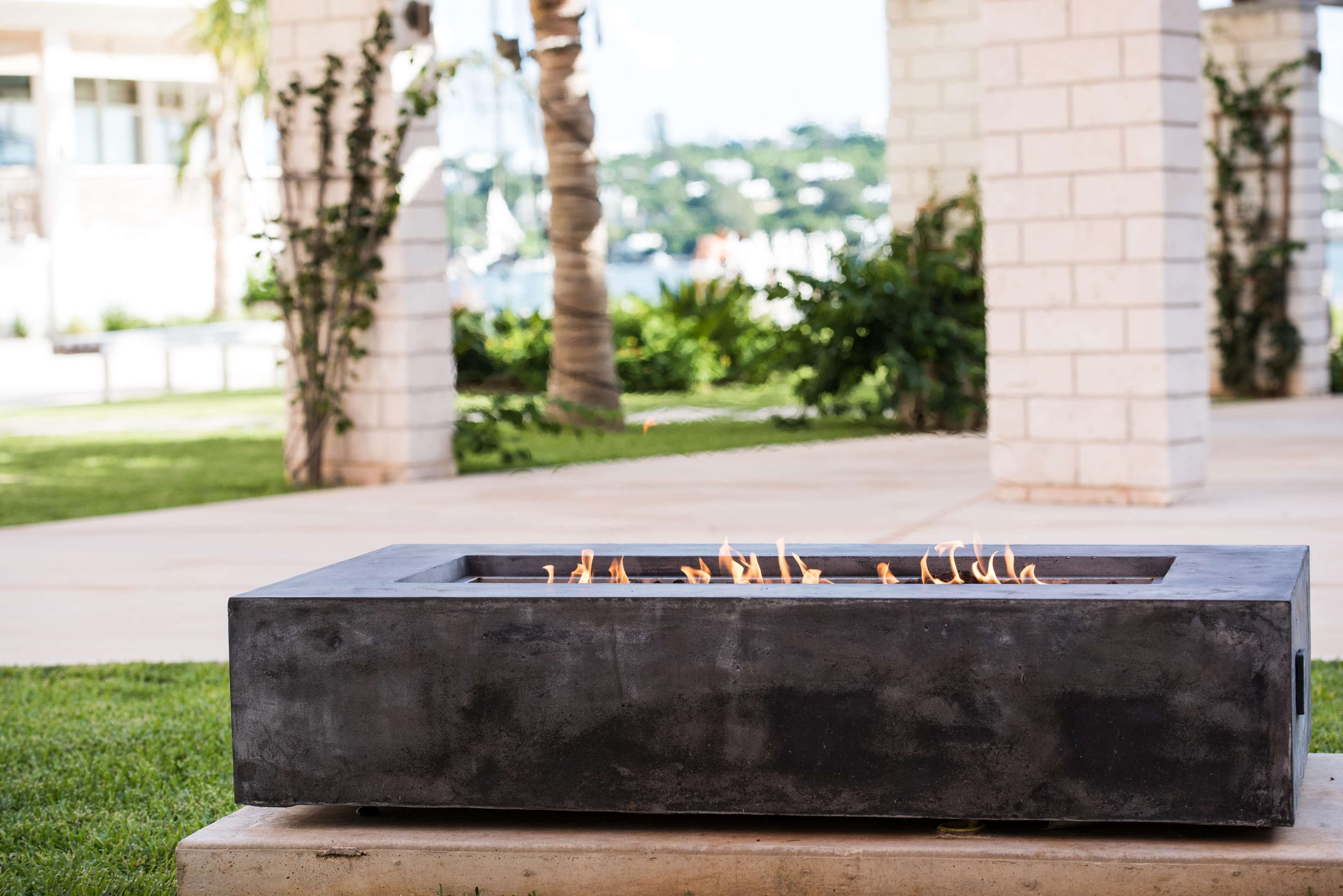 Una estructura rectangular para fuego al aire libre con llamas, ubicada sobre una base de concreto, rodeada de follaje y columnas arquitectónicas.