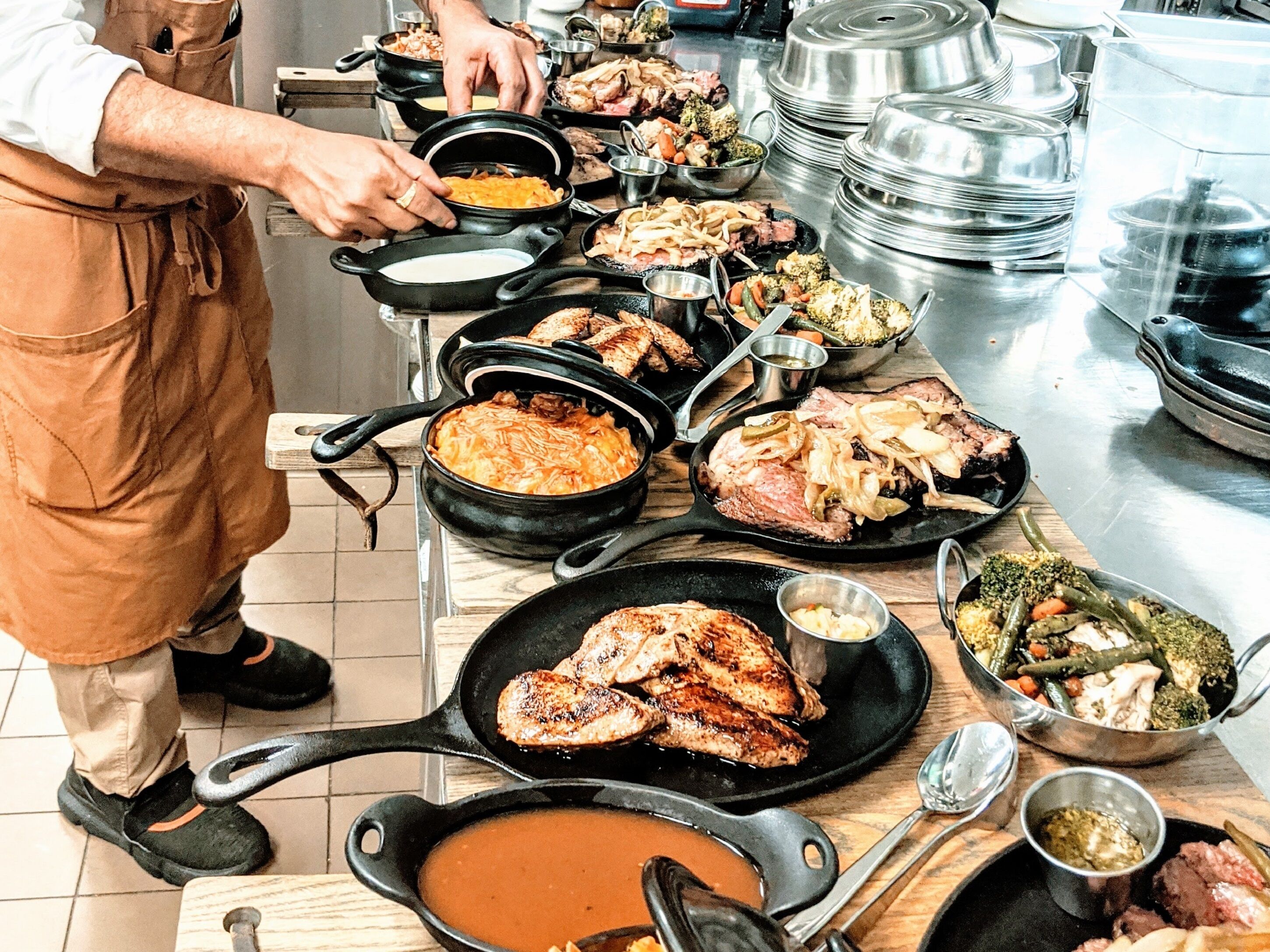 A kitchen with various cooking pots and pans on the counter, a person in an apron, and a variety of dishes being prepared.