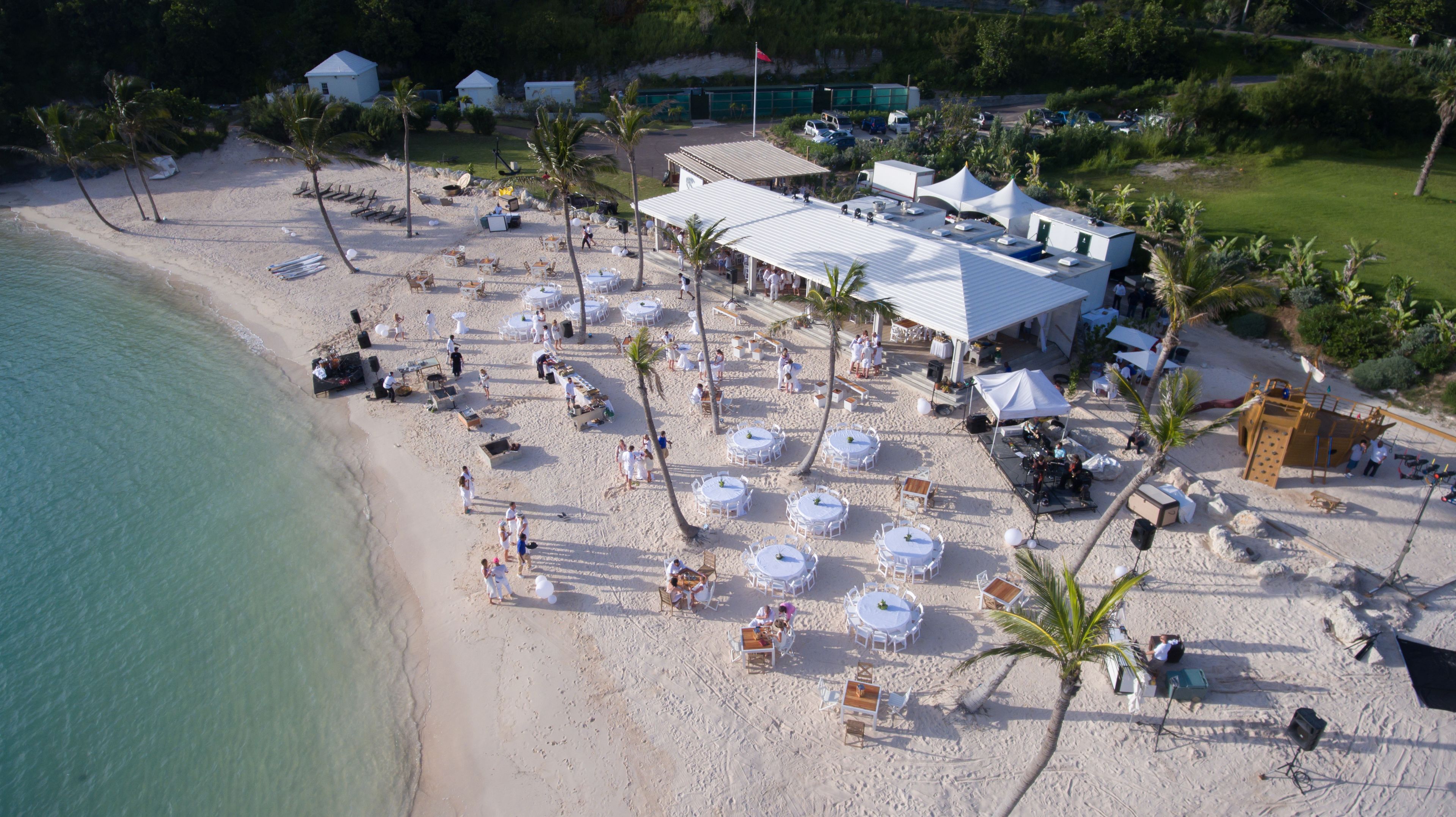 An aerial view of a beachside resort with multiple white tents, palm trees, and a sandy beach.
