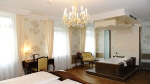 Ein Hotelzimmer mit einem großen Bett, einer Sitzecke mit einem Sessel, einem Schreibtisch, einem Kronleuchter und einem separaten Badezimmer mit Badewanne und Dusche.
