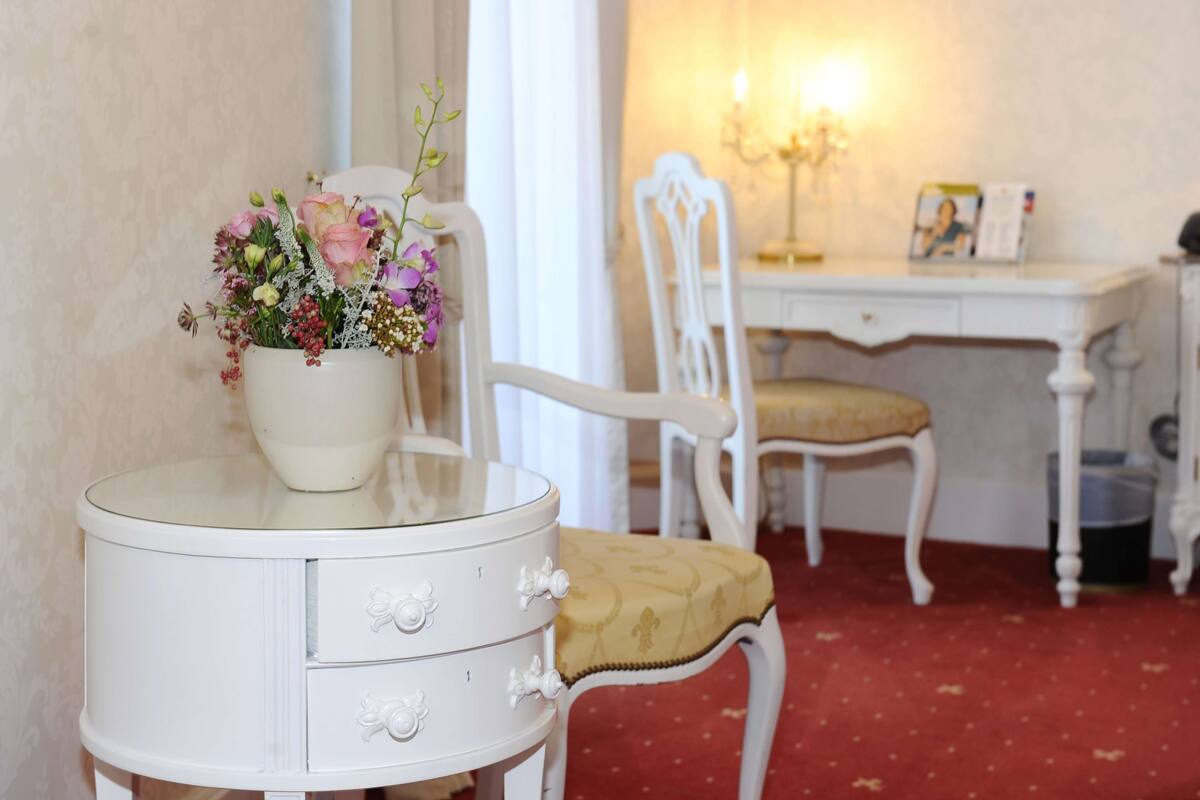 Un tocador blanco con cubierta de vidrio y un florero. Una silla blanca con cojín amarillo se encuentra frente a un escritorio blanco con un marco de fotos y un candelabro.