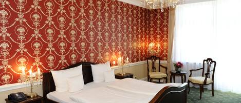 A neatly made bed with white linens, a chandelier, and a patterned wallpaper.