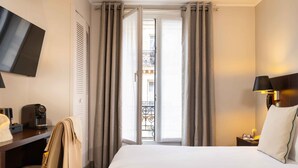 Quarto de hotel com cama, escrivaninha com cafeteira, cadeira e televisão fixada na parede.