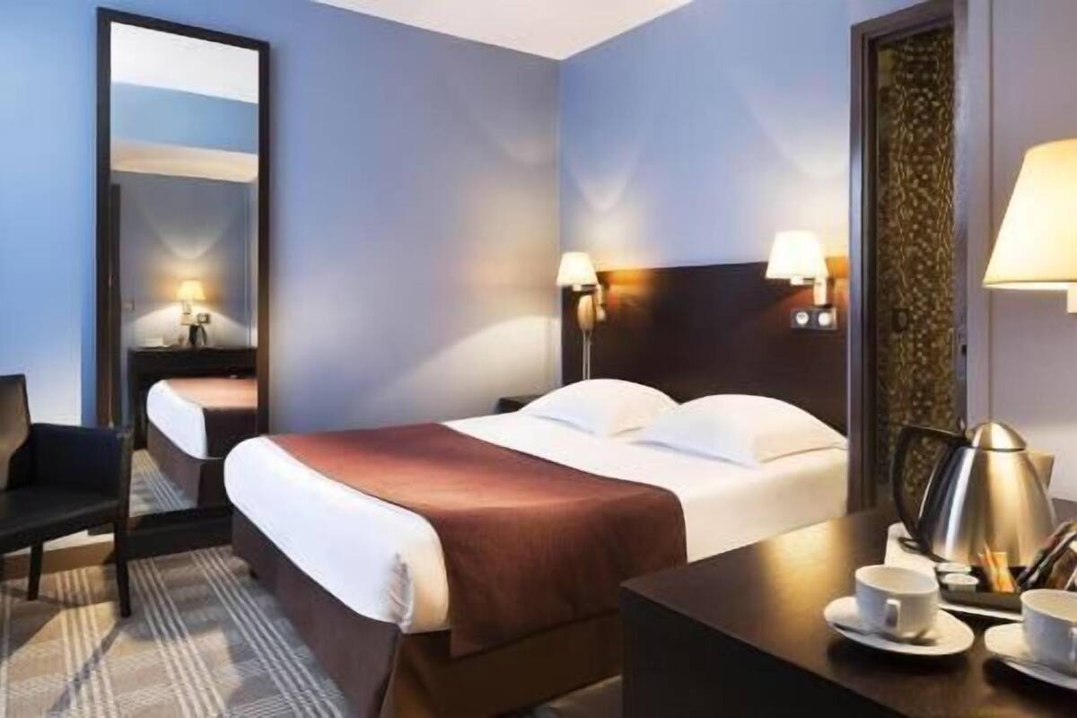 A hotel room with a bed, bedside lamps, a desk with a cup and saucer, and a chair.