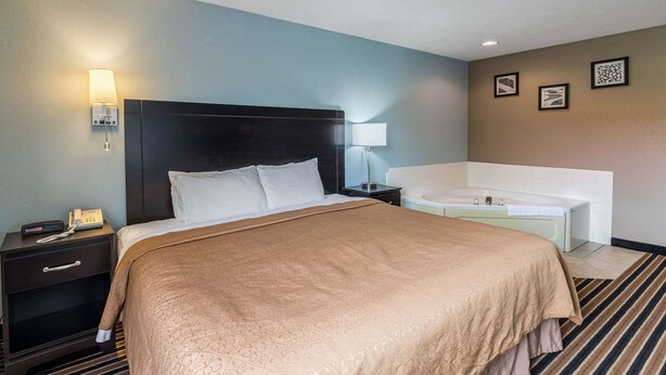 Habitación de hotel con una cama grande, una mesita de noche con lámpara, una mesita auxiliar y una bañera.