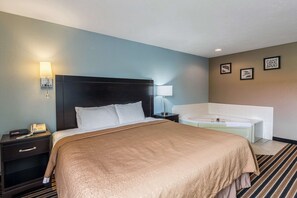 A hotel room with a large bed, a bedside table with a lamp, a nightstand, and a bathtub.