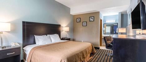 A hotel room with a bed, bedside table, lamp, and a desk with a chair.
