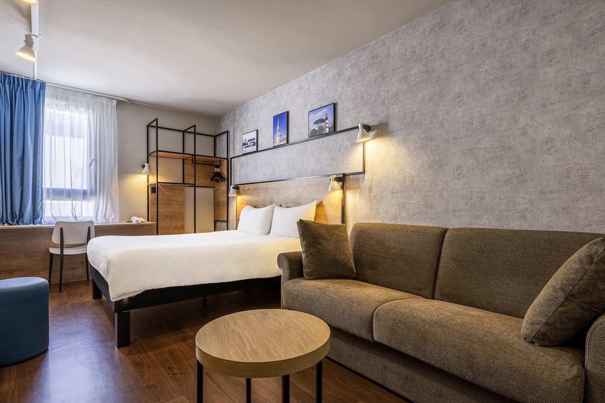 A modern hotel room with a bed, sofa, round table, and a desk with a chair.