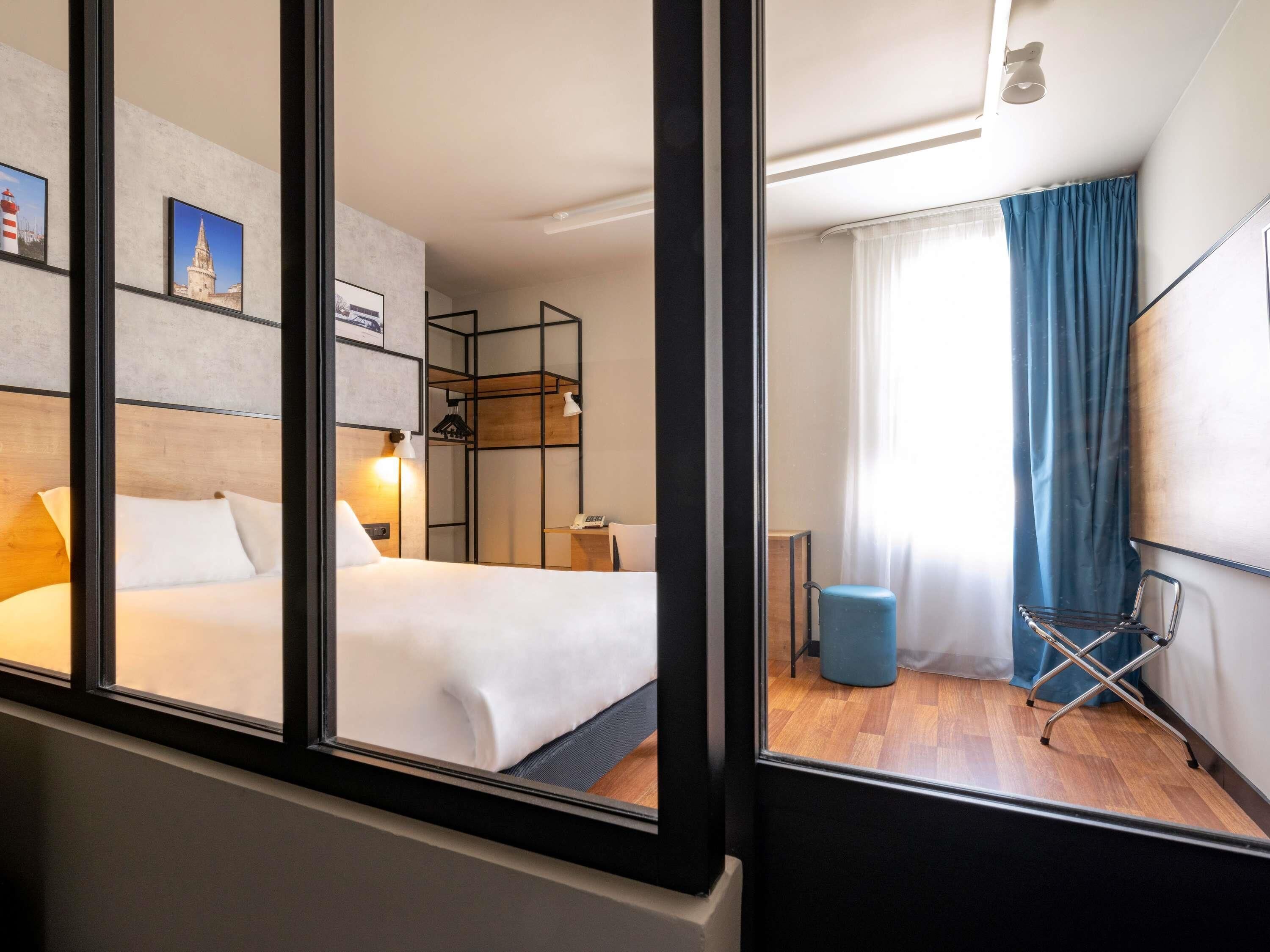 Habitación de hotel con cama, mesita de noche, lámpara y vista a la ciudad a través de una ventana.