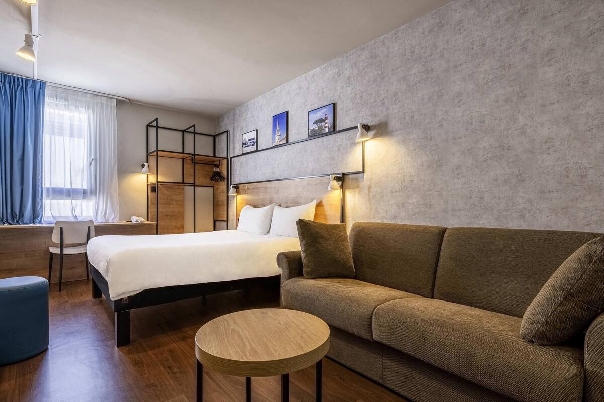 A modern hotel room with a bed, sofa, round table, and a desk with a chair.