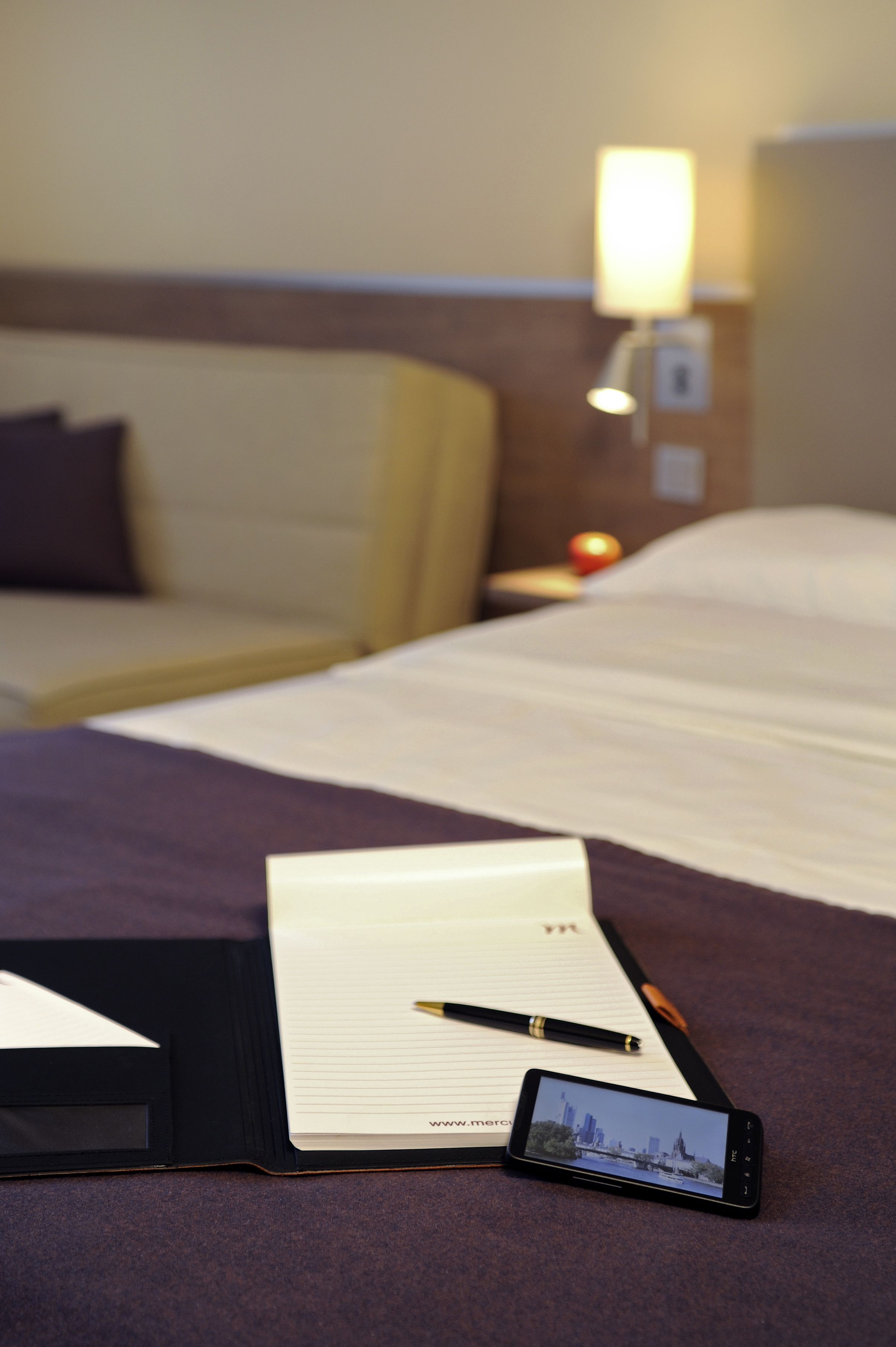A neatly made bed with a pillow, a bedside table with a lamp, and a smartphone displaying a cityscape.