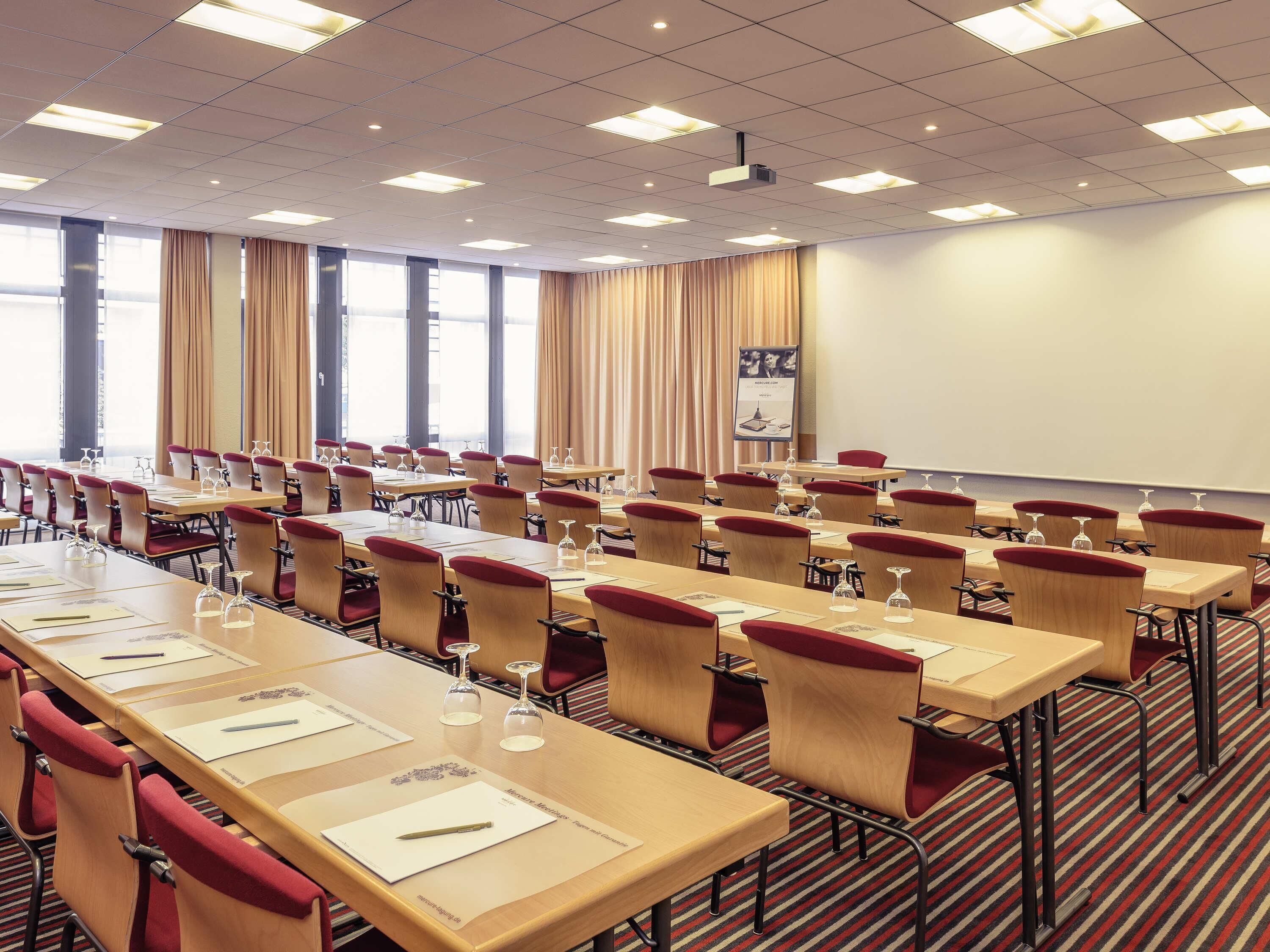 Ein Konferenzraum mit mehreren Tischen und Stühlen, einer Projektionsleinwand und einem großen Fenster mit Vorhängen.