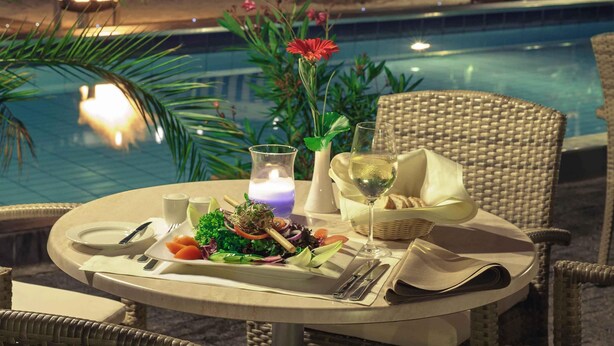 A dining table set with a salad, wine glass, and candle, overlooking a pool area with lounge chairs and a tiki lamp. - Mercure Hotel Frankfurt Eschborn Ost (Eschborn)