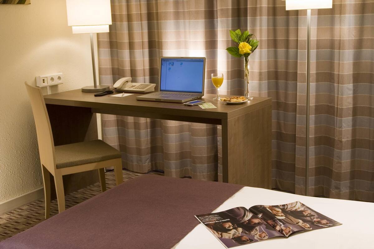 A hotel room with a desk, laptop, phone, and a magazine on the bed.