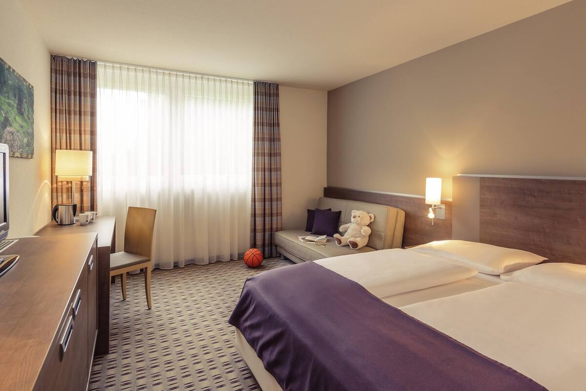 A hotel room with a bed, a desk, a chair, a lamp, and a teddy bear.