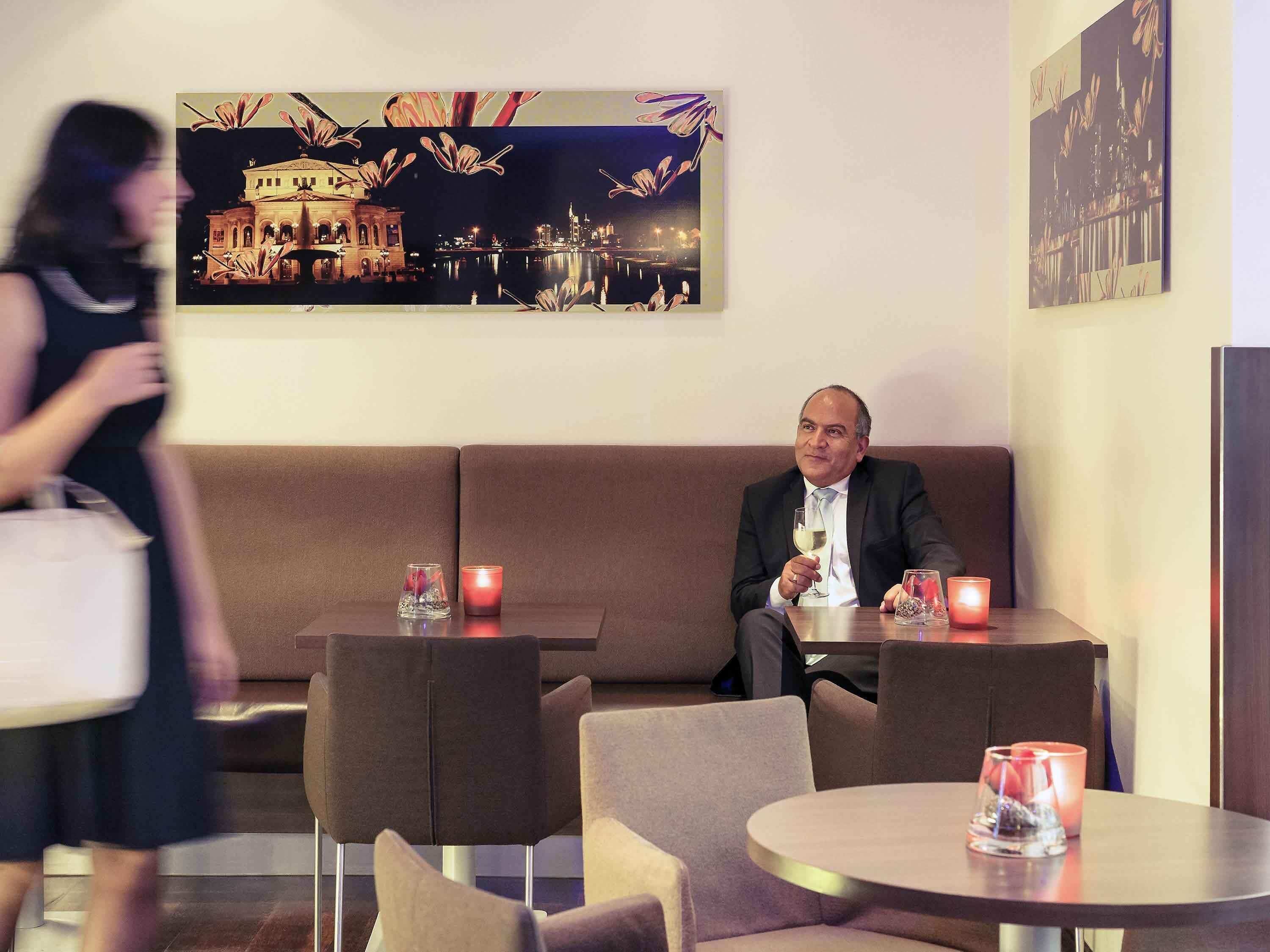 A man seated at a table with a glass of wine, a sofa, and a painting of a cityscape in the background.