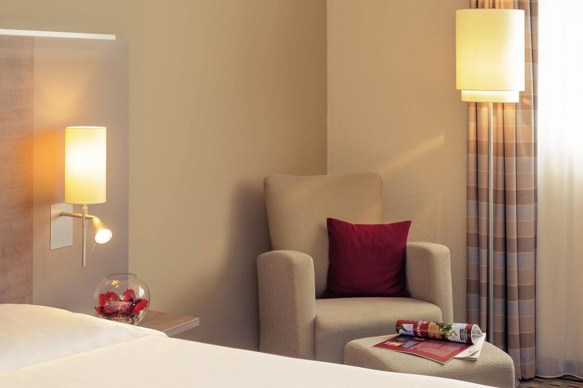 A hotel room with a bed, a chair with a red pillow, a nightstand with a lamp, and a glass of water.