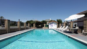 A rectangular swimming pool with clear blue water, surrounded by a stone wall and a fence. There are lounge chairs and a small table with an umbrella on one side.