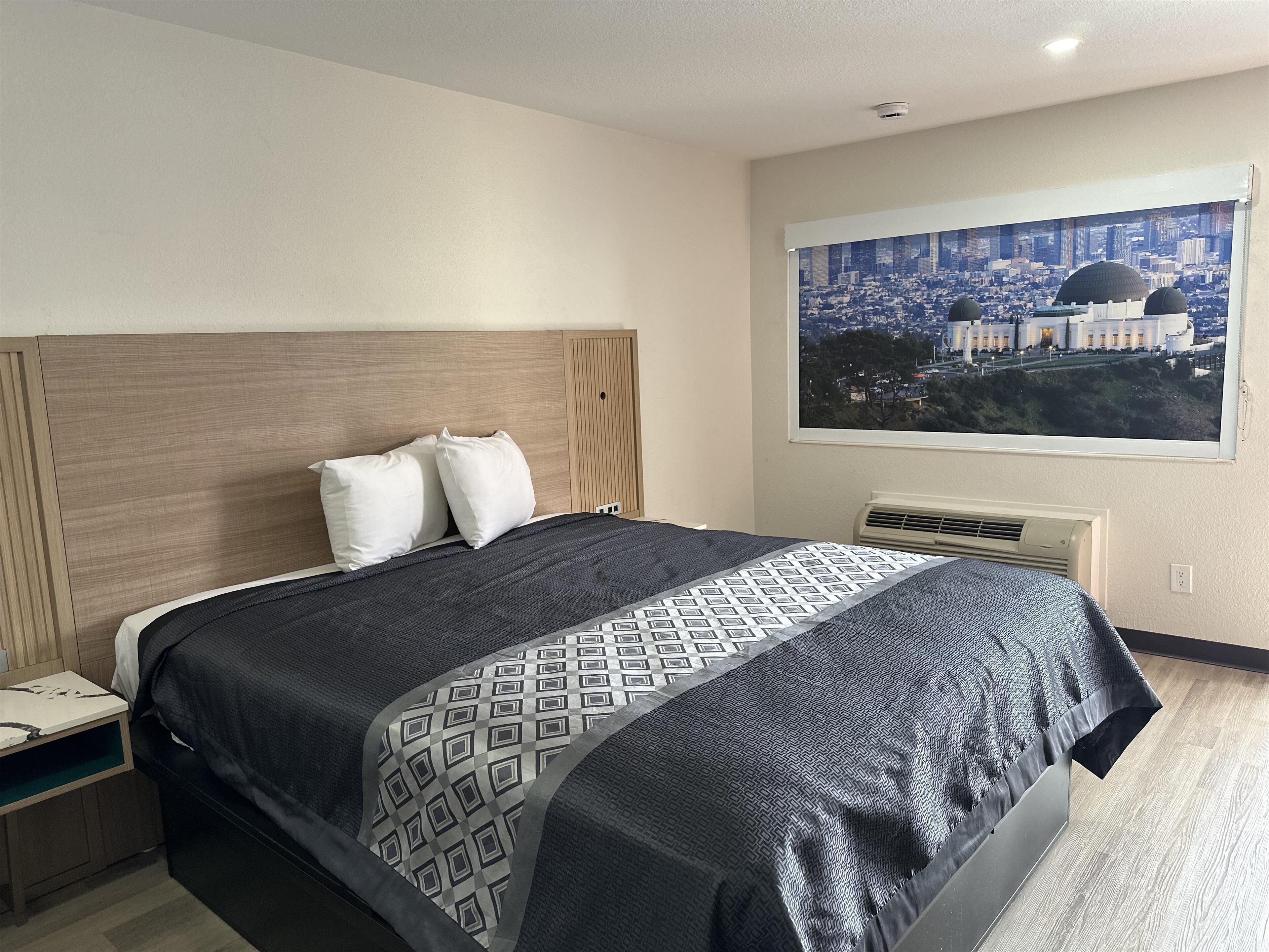 A hotel room with a bed, a nightstand, a window with a cityscape view, and a wall-mounted air conditioner.