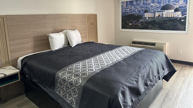 A hotel room with a bed, a nightstand, a window with a cityscape view, and an air conditioning unit.