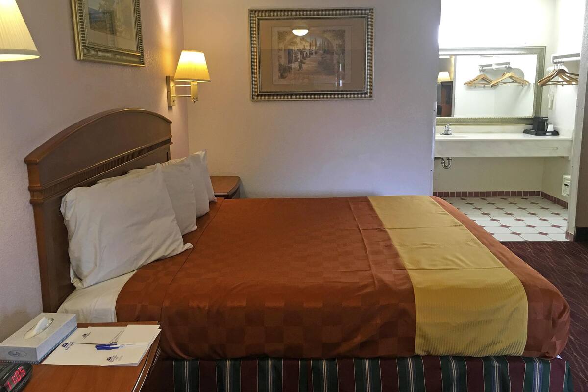 Une chambre d’hôtel avec un lit, une table de chevet avec une lampe, un tableau encadré au mur et une salle de bain visible à travers une porte ouverte.
