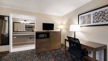 A hotel room with a bed, a desk with a chair, a television, a lamp, and a bathroom with a sink and mirror.
