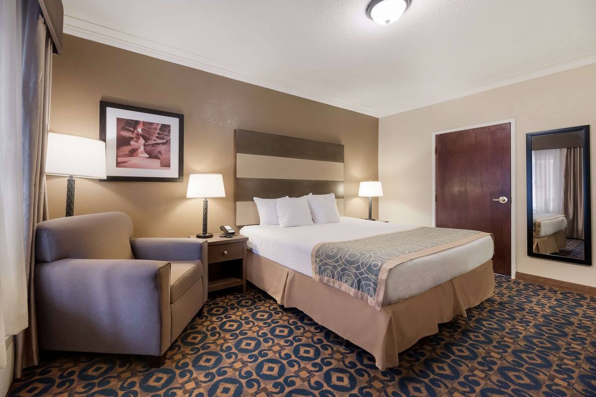 A hotel room with a bed, a chair, a nightstand, a lamp, a framed picture on the wall, and a patterned carpet.
