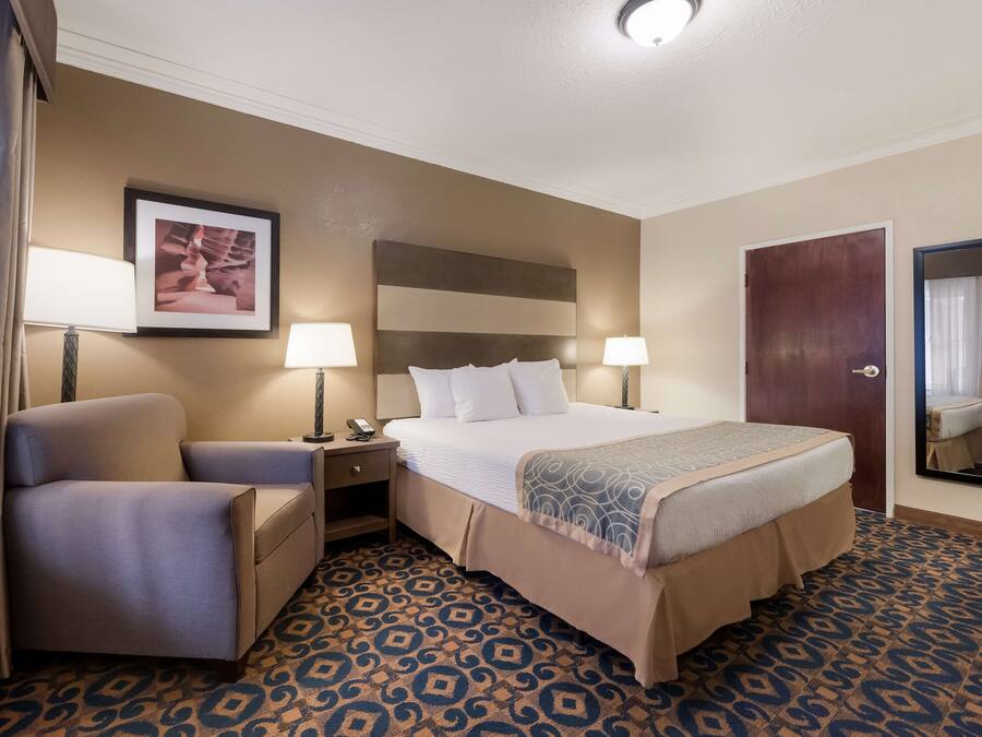 A hotel room with a bed, a chair, a nightstand, a lamp, a framed picture on the wall, and a patterned carpet.