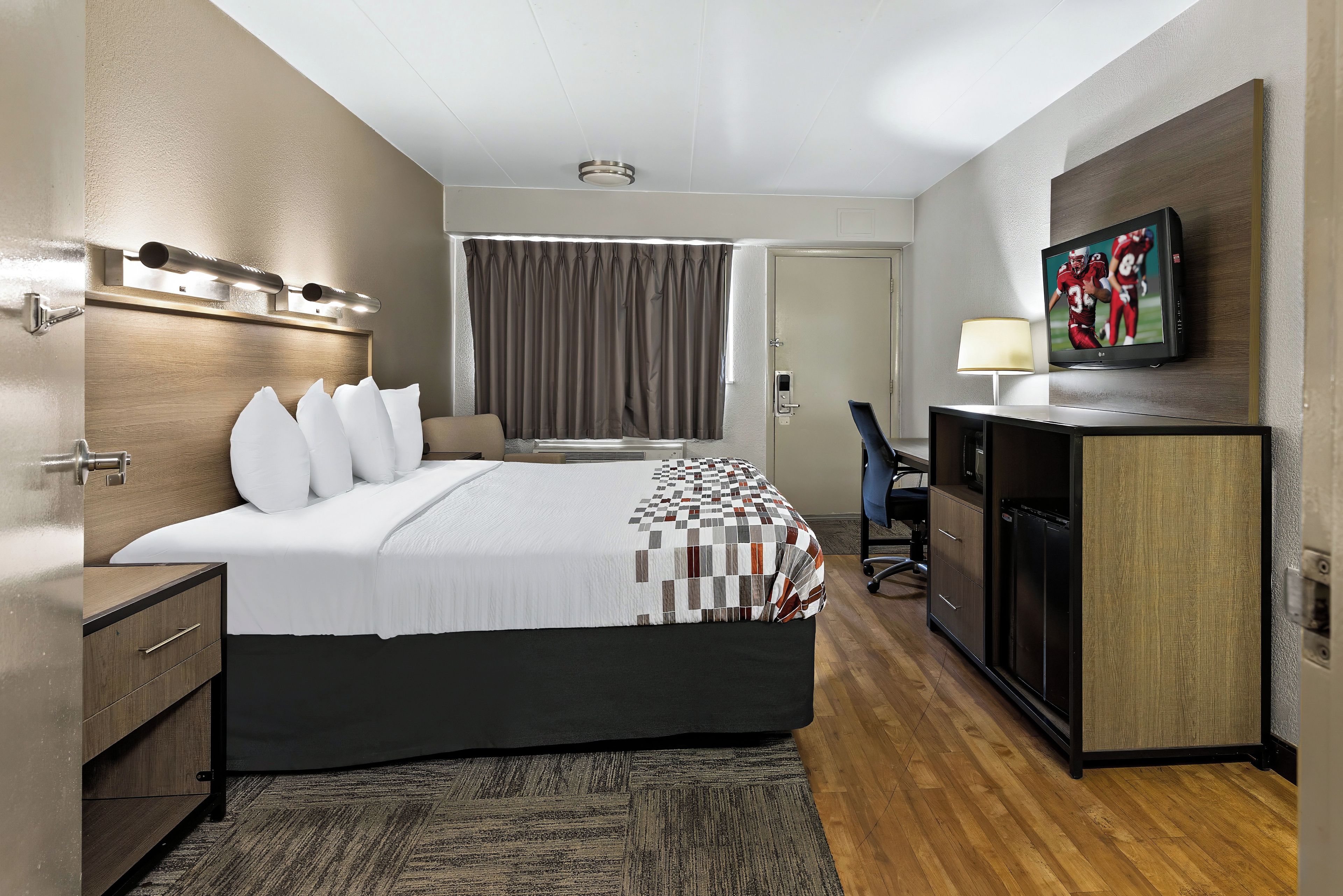 A hotel room with a bed, bedside table, lamp, TV, and a desk with a chair.