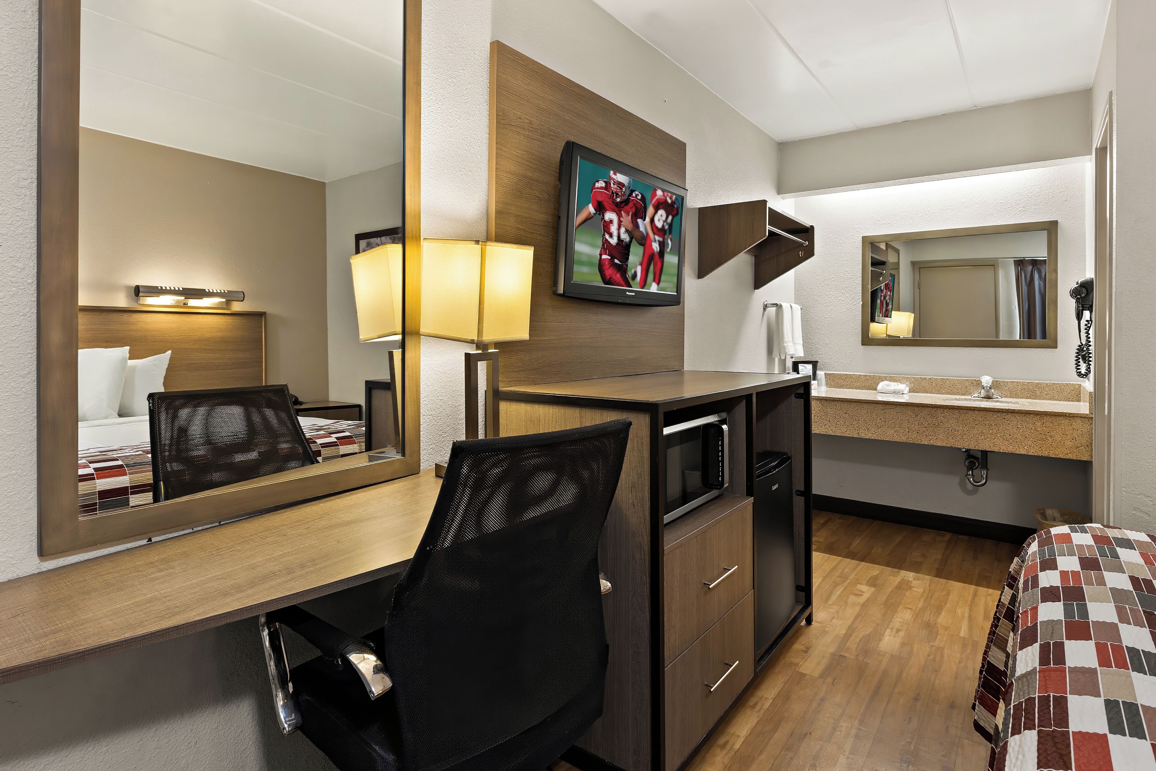 A hotel room with a wooden desk, a black office chair, a television mounted on the wall, a microwave, a small refrigerator, a bed with a checkered bedspread, a bedside table, and a mirror.