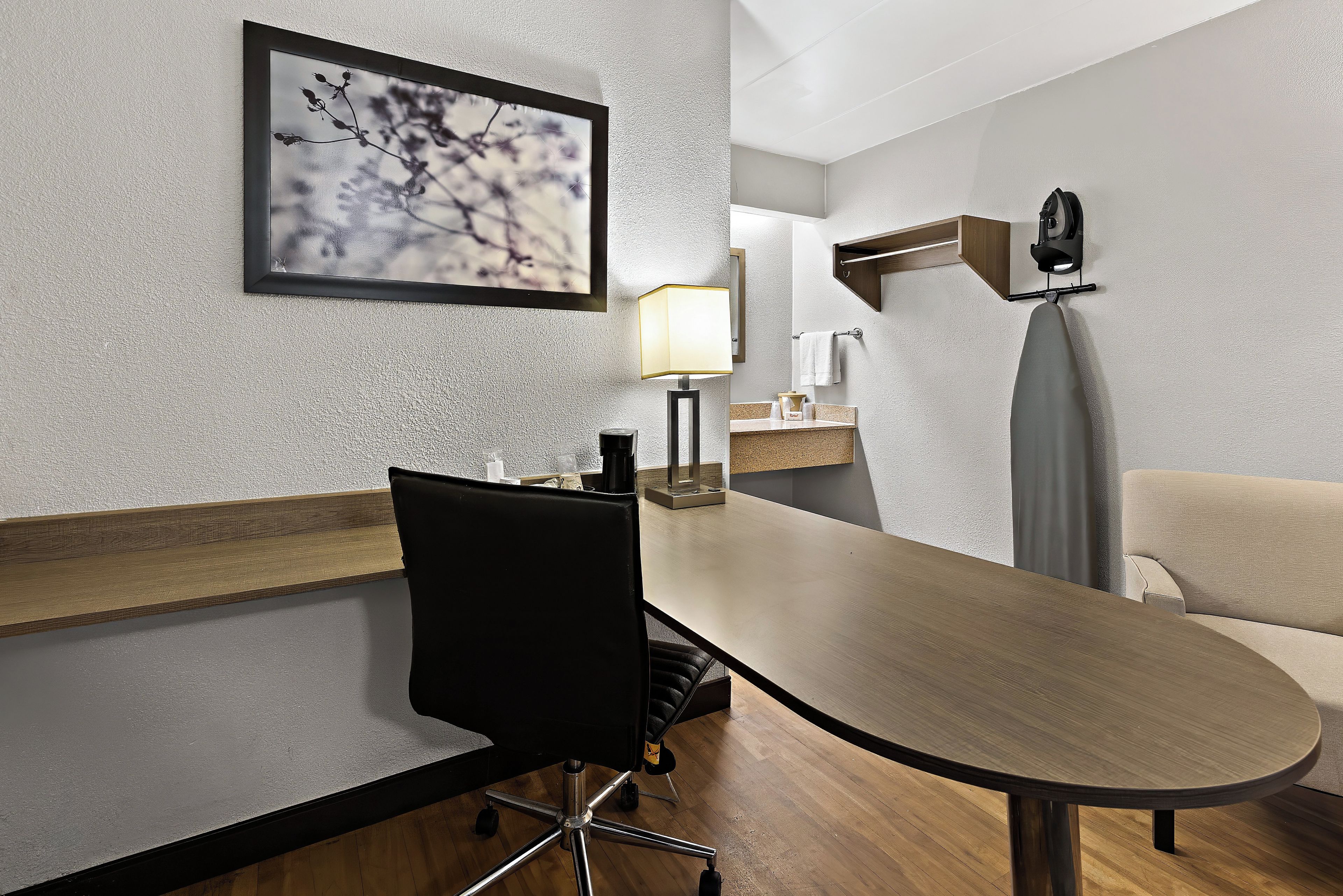 Habitación de hotel con una larga mesa de madera, una silla de oficina negra, un cuadro enmarcado en la pared, una lámpara de noche y una mesita de noche con artículos de aseo.