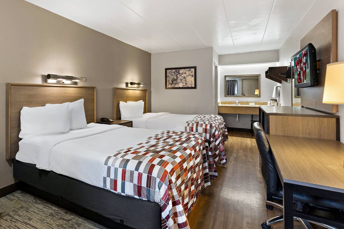 A hotel room with two beds, a desk, a chair, a TV, and a bathroom with a mirror and sink.