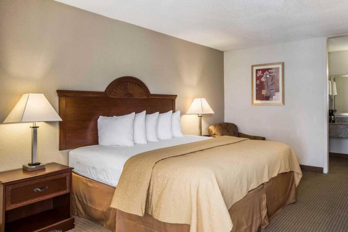 A hotel room with a bed, two bedside lamps, a nightstand, a framed picture on the wall, and a bathroom door.