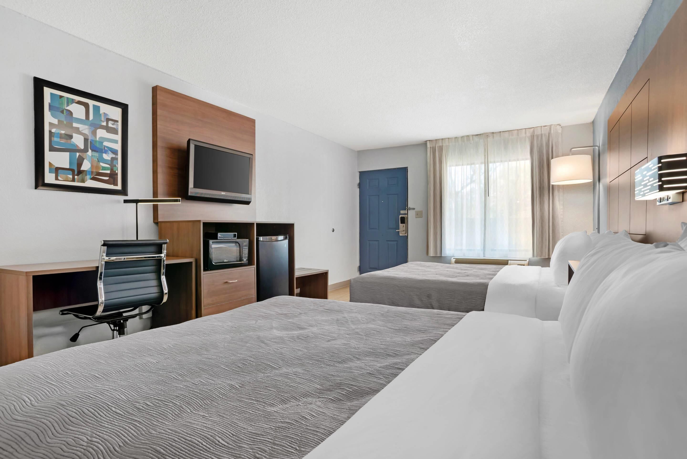 A hotel room with two beds, a desk with a chair, a television, a microwave, and a door with a blue frame.