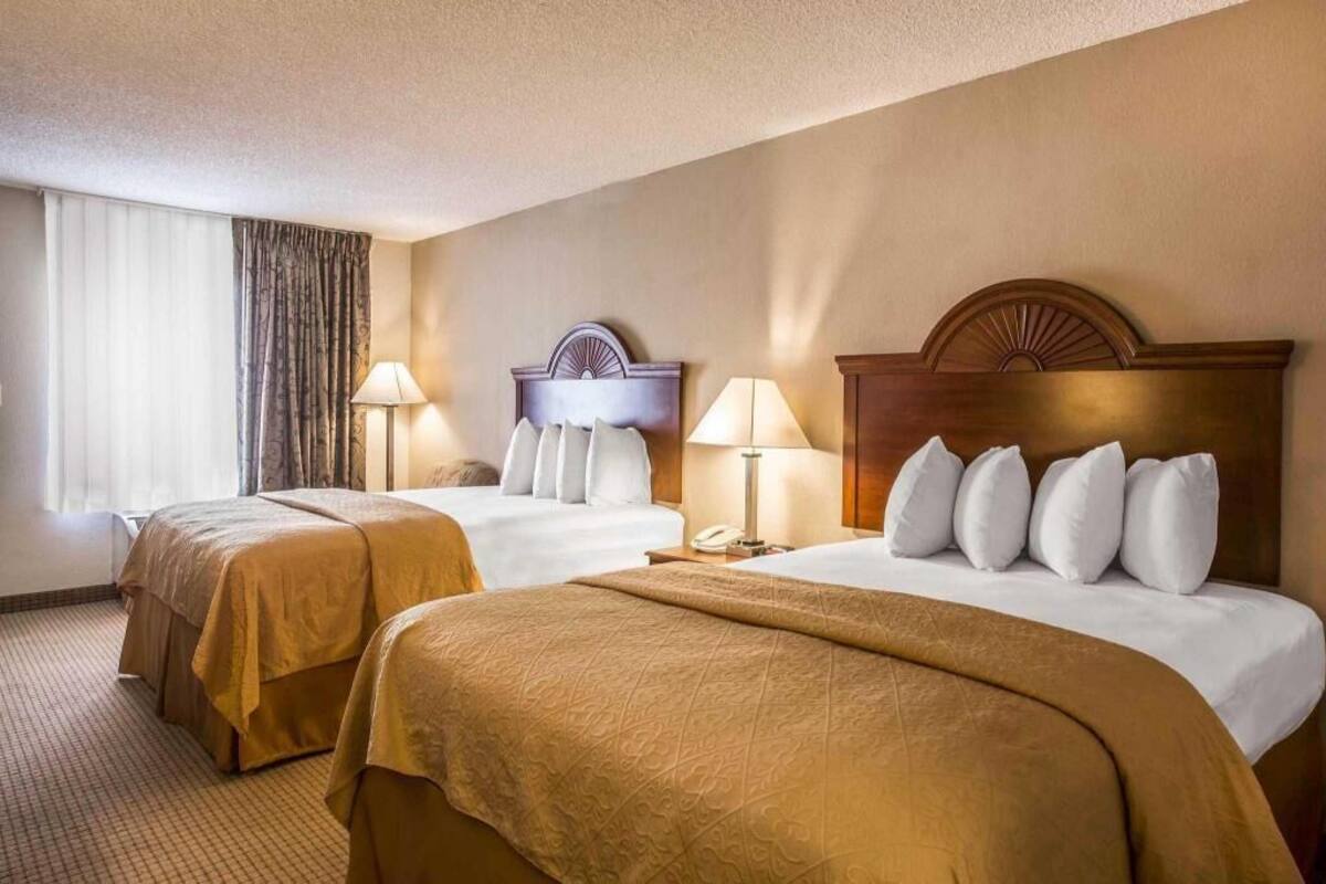 A hotel room with two beds, a large window with curtains, a headboard with a decorative design, and a nightstand with a lamp.