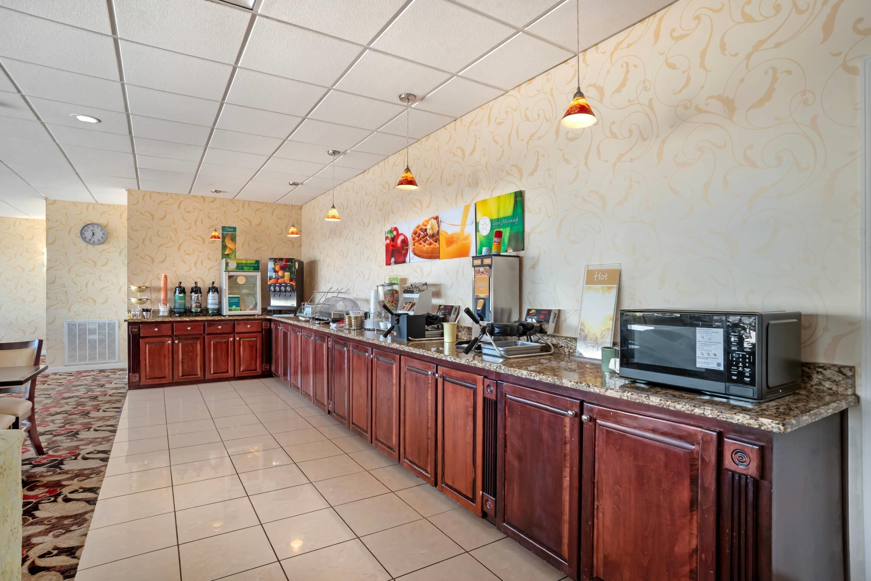 A spacious kitchen with a long counter, wooden cabinets, and a microwave. There are various appliances and a clock on the wall.