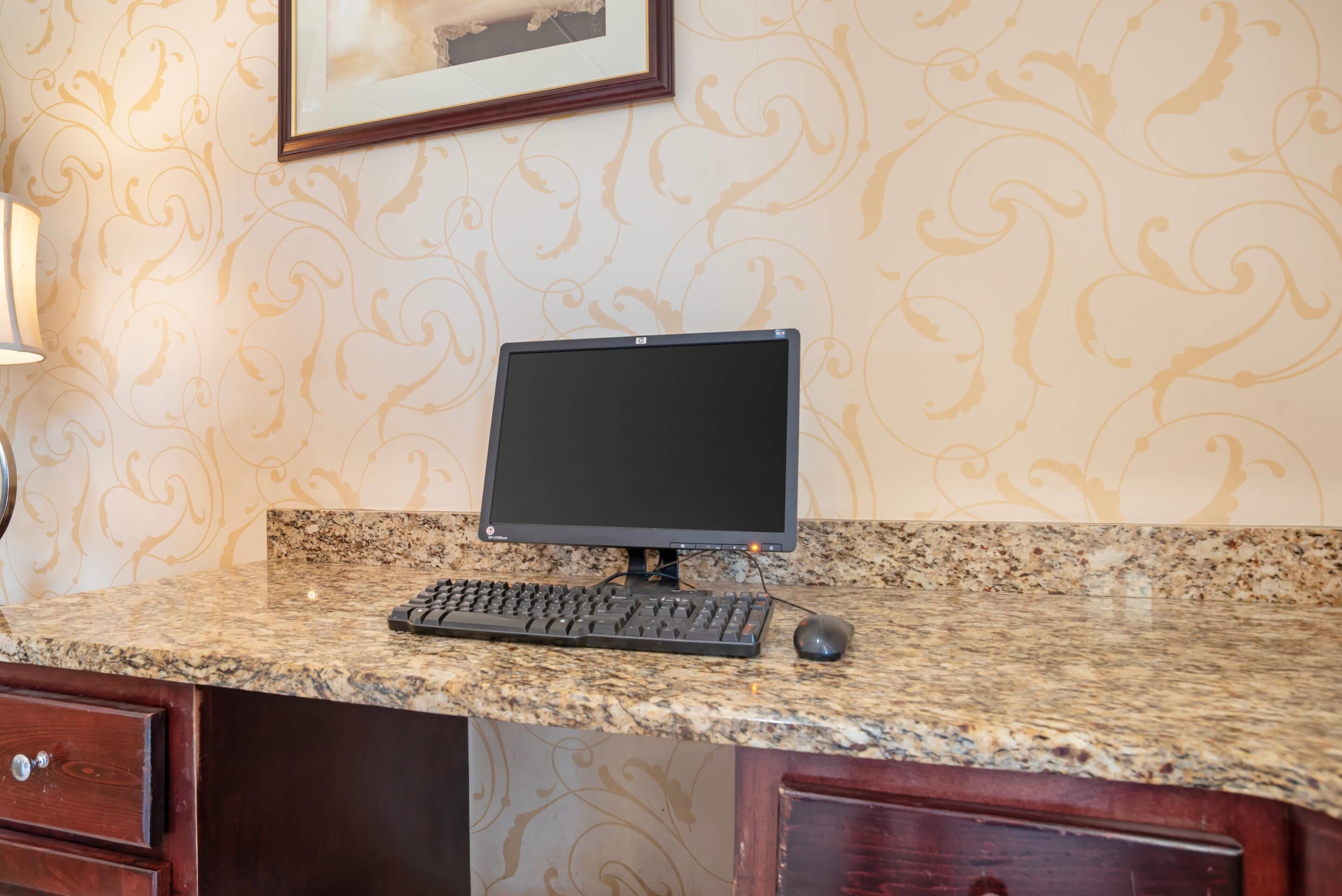 A desktop computer setup on a granite countertop with a patterned wallpaper, a framed picture on the wall, and a wooden cabinet below.