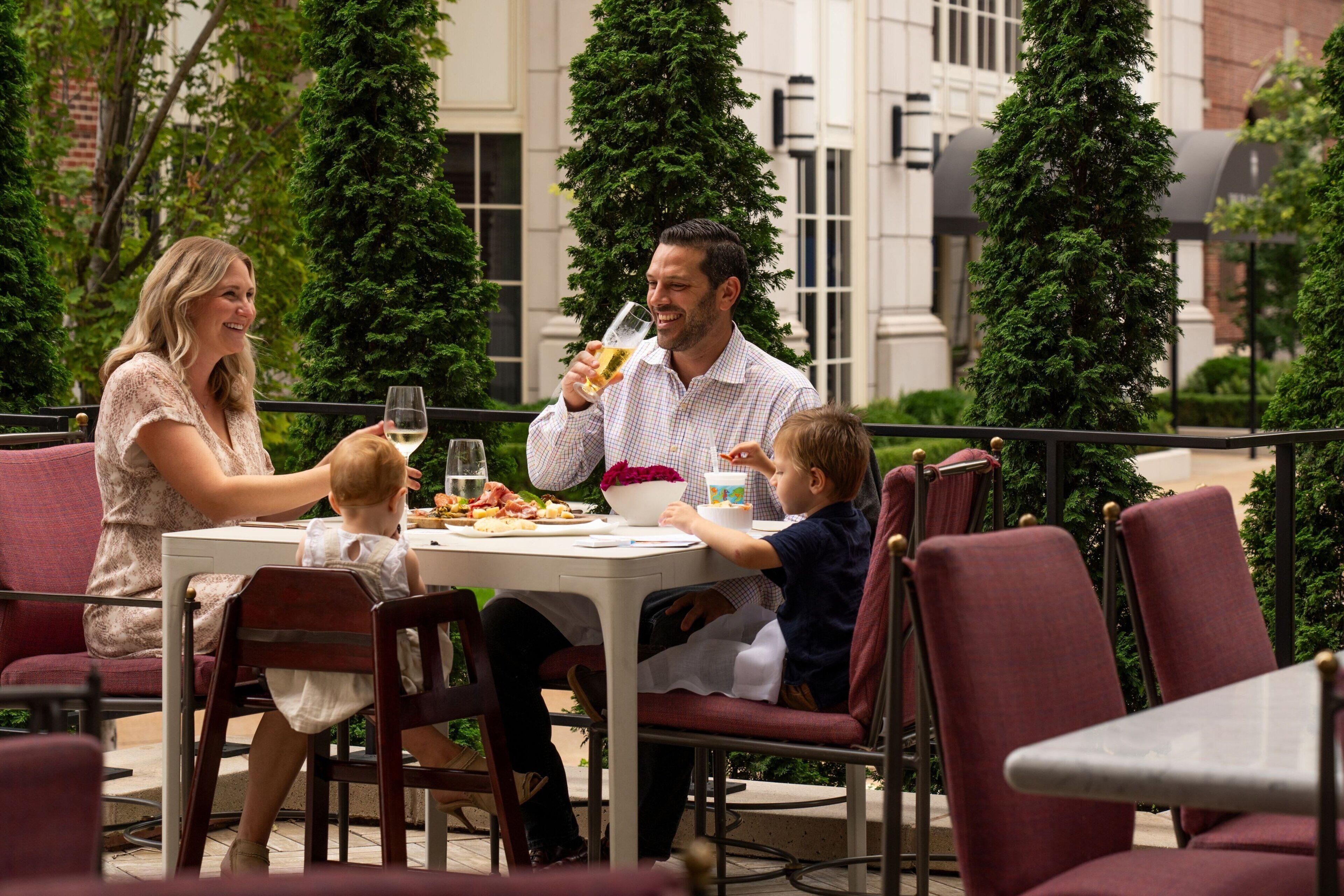 A family dining outdoors at a restaurant with a white table, red chairs, and a brick building in the background.