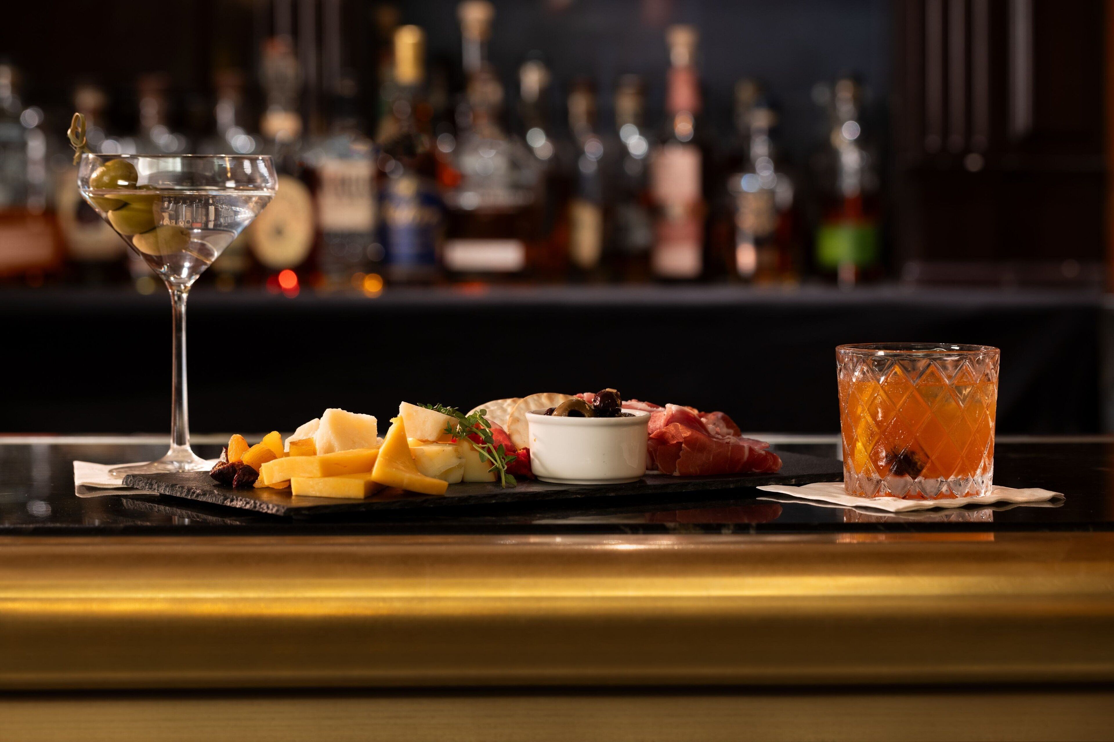 A bar setting with a martini glass, a cocktail glass, and a cheese board with various cheeses and accompaniments.