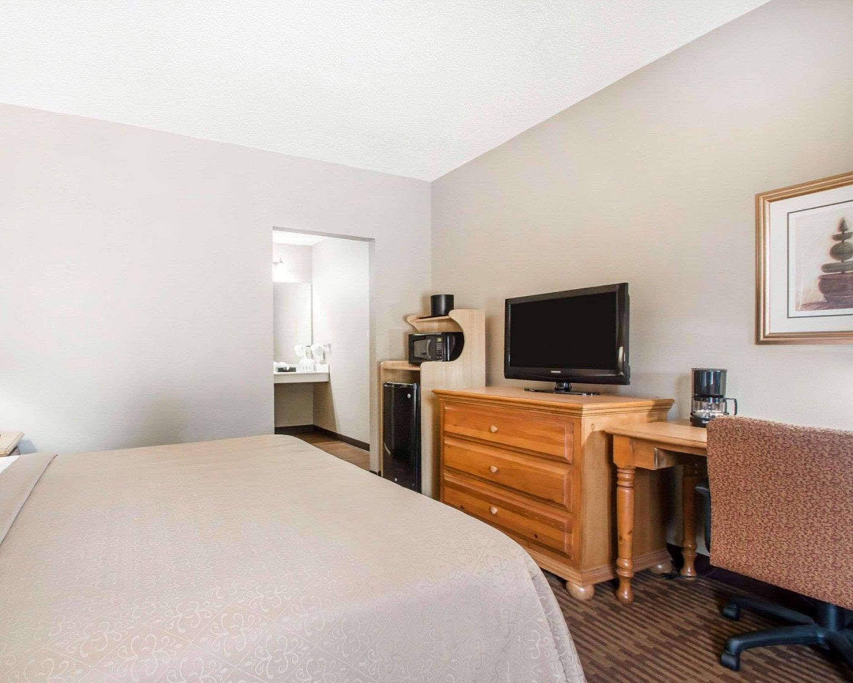 Ein Hotelzimmer mit einem Bett, einem Schreibtisch mit Stuhl, einem Fernseher, einer Mikrowelle, einer Kaffeemaschine und einem gerahmten Bild an der Wand.
