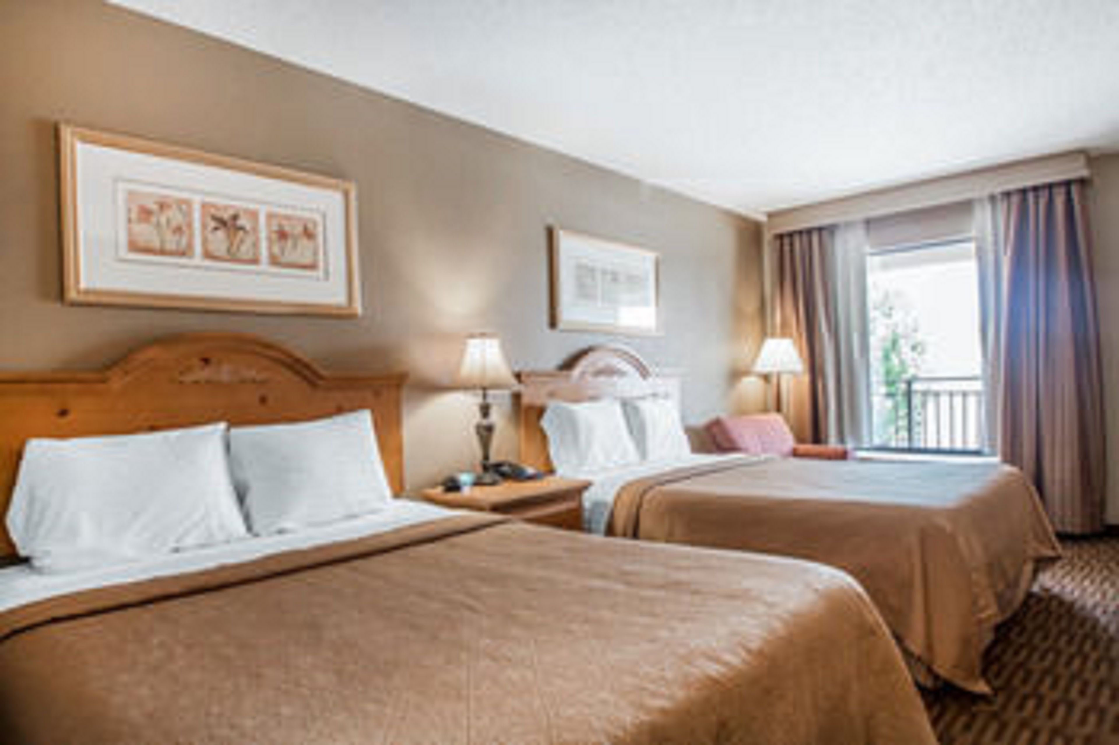 A hotel room with two beds, a wooden headboard, bedside tables with lamps, framed artwork on the wall, and a window with curtains.